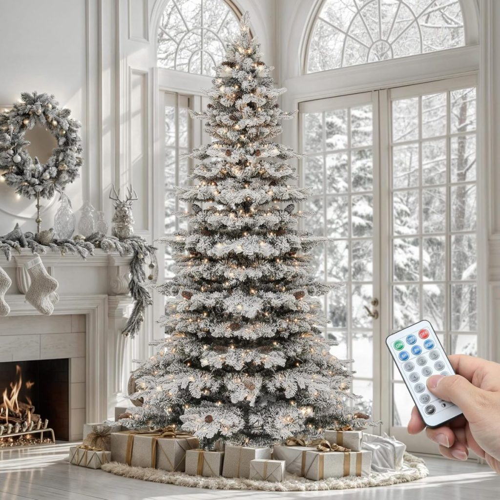 A hand holds a remote control in front of a decorated, flocked Christmas tree with lights in a snowy, elegant living room with wrapped gifts and a lit fireplace.
