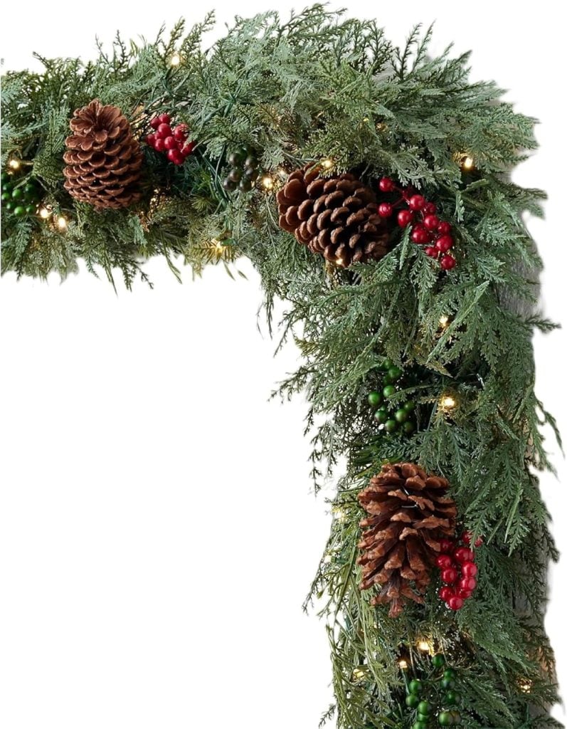 A green holiday garland decorated with pine cones, red berries, and small white string lights.