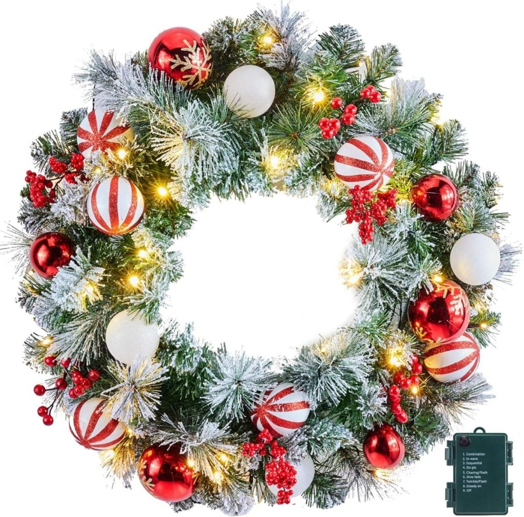 A decorated Christmas wreath with red, white, and gold ornaments, artificial snow, berries, warm white lights, and a small black battery pack is perfect as festive window art.