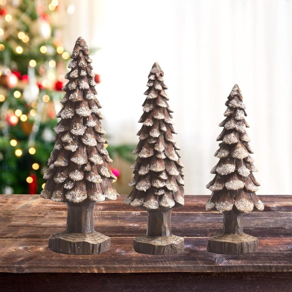 Three wooden pine tree decorations of varying heights sit on a wooden surface, with a blurred decorated Christmas tree in the background.