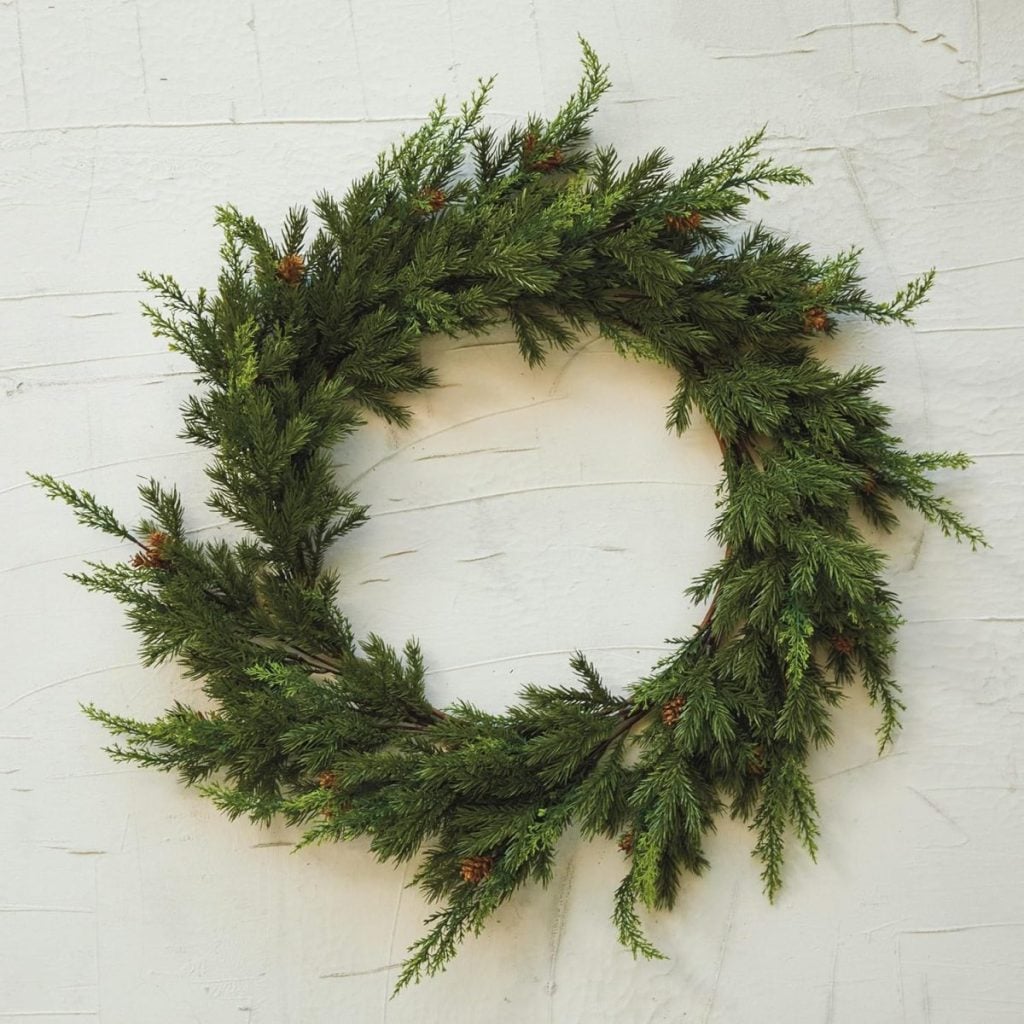 A simple green wreath made of pine branches with small brown pinecones hangs on a white textured wall, adding a touch of window art charm to the decor.