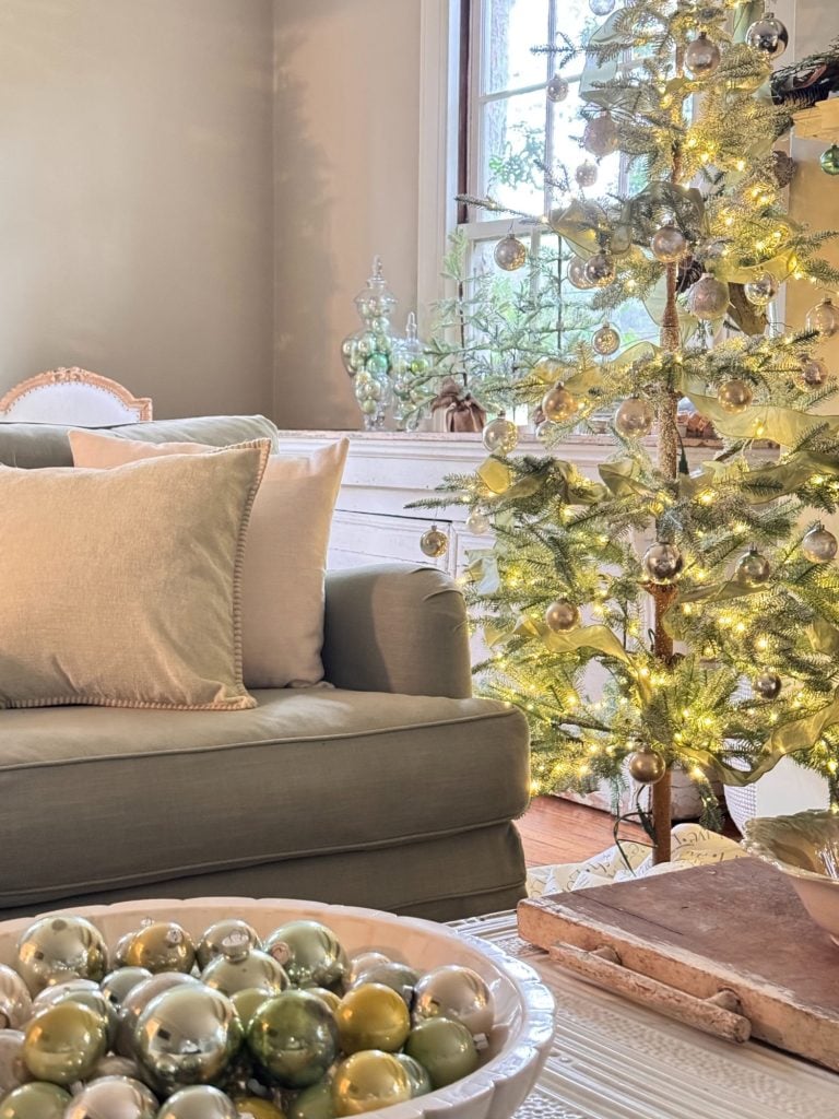Living room with a lit Christmas tree decorated with silver and gold ornaments, a green sofa, and a bowl of ornaments on a white coffee table.