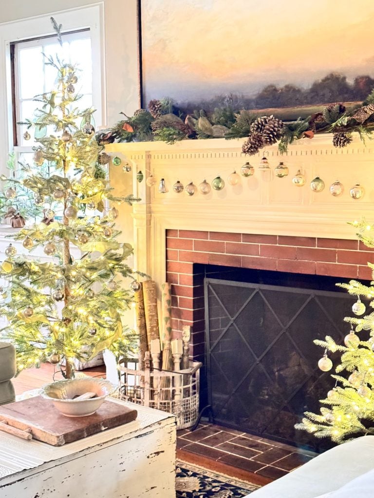 A living room with a decorated mantle, lit Christmas trees, hanging ornaments, pinecones, and a basket of firewood by a fireplace.