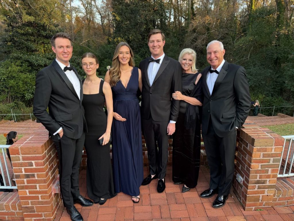 Six adults dressed in formal evening wear stand together outdoors on a brick patio, smiling at the camera with trees in the background.