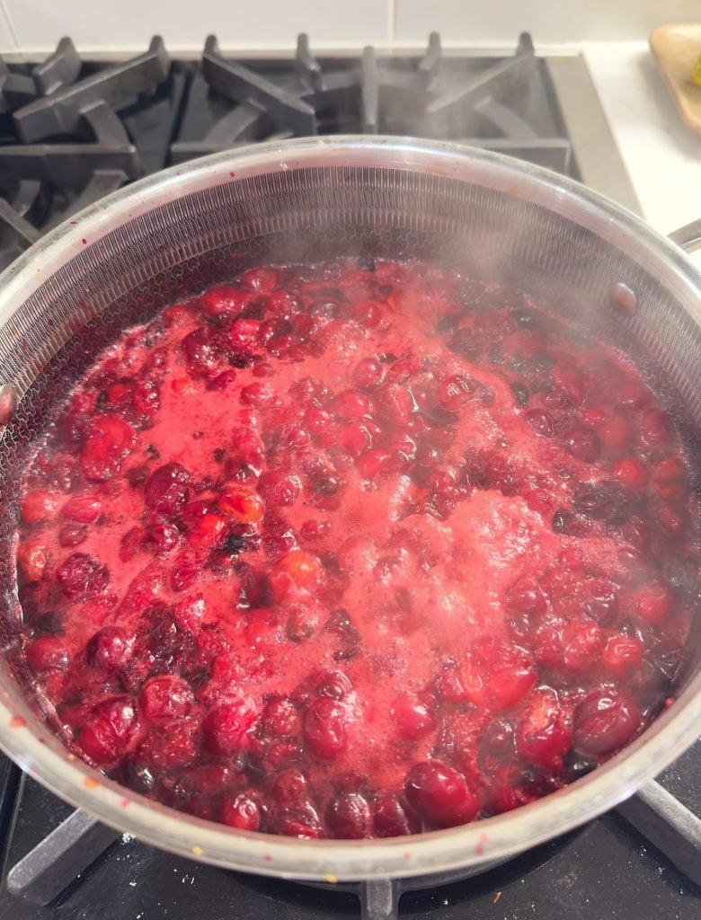 A saucepan of cranberries cooking on a stovetop, with red foam forming on the surface and steam rising.