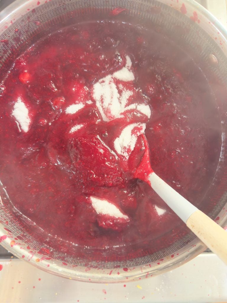 A pot of red berry mixture is being stirred with a spatula, with visible sugar sprinkled on top and steam rising from the hot contents.