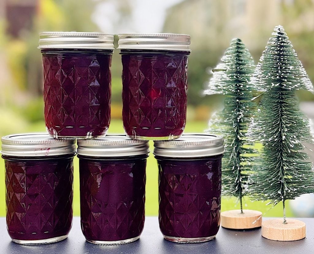 Five glass jars filled with dark purple jam are stacked in a pyramid next to two small artificial pine trees.