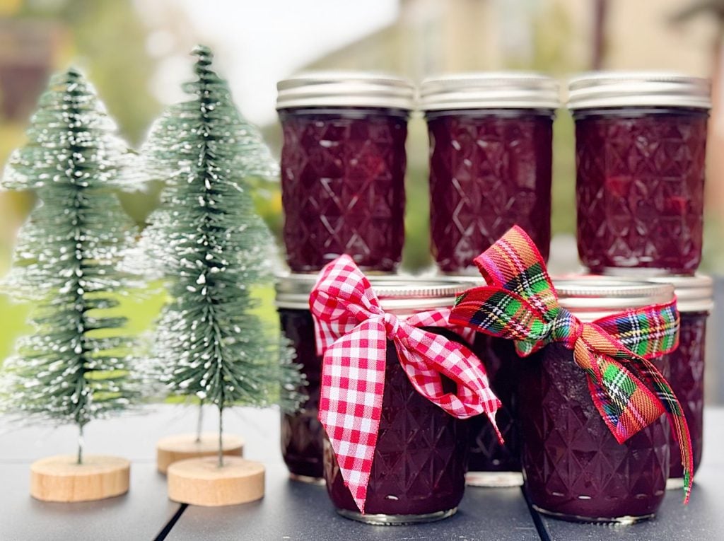 Six jars of jam are stacked next to two miniature Christmas trees; two jars have festive ribbon bows tied around them.