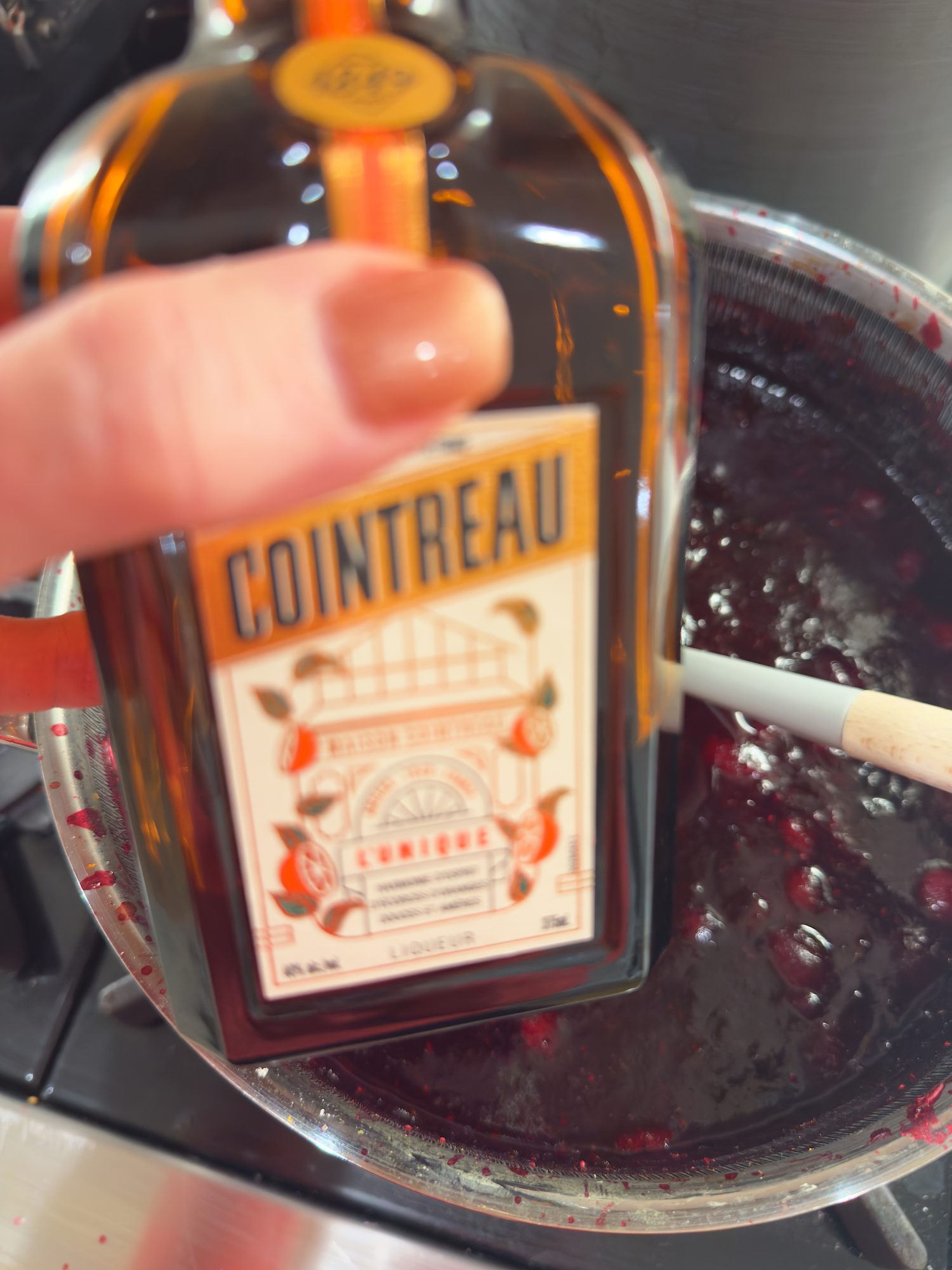 A hand holds a bottle of Cointreau liqueur above a saucepan filled with a dark, berry mixture being stirred on a stove.