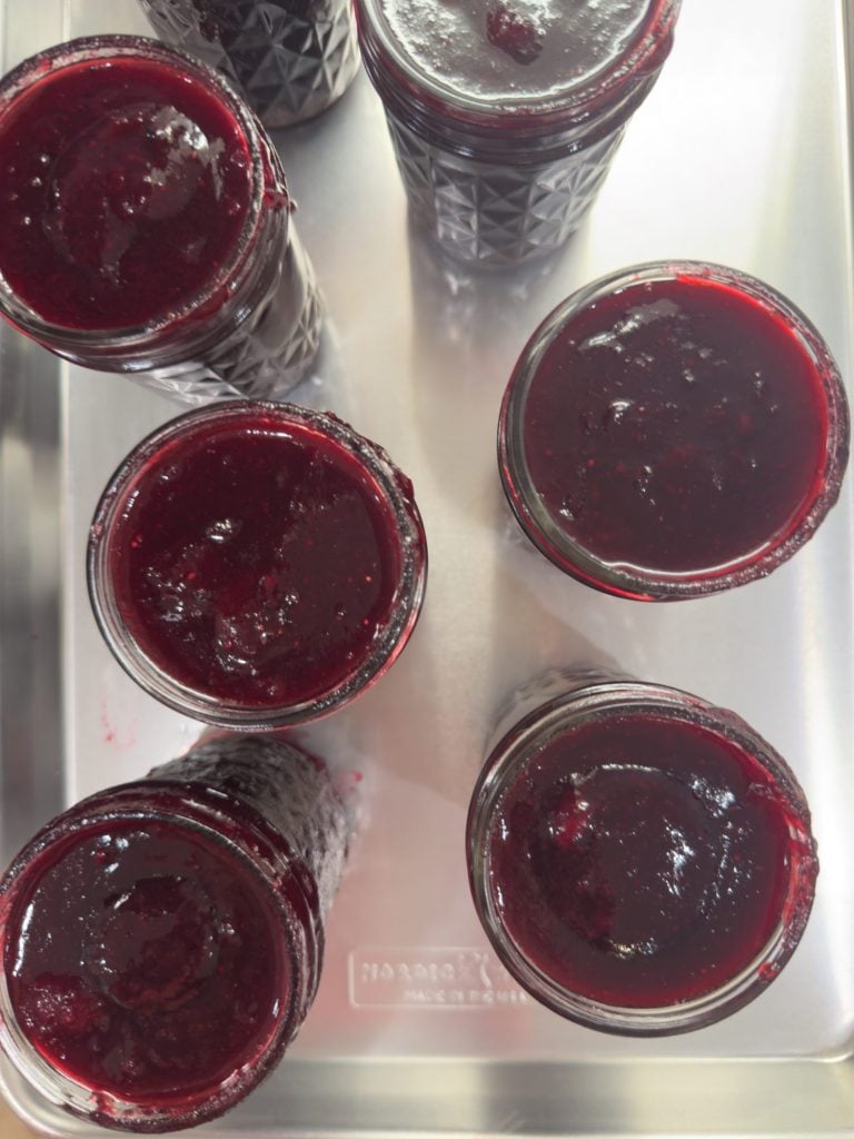 Six glass jars filled with dark red jam are arranged on a metal tray, viewed from above. Some jars have small spills around the rim.