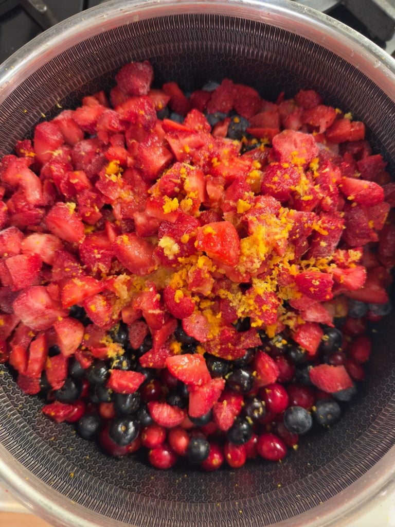 A metal colander filled with chopped strawberries, blueberries, cranberries, and a sprinkle of grated orange zest.
