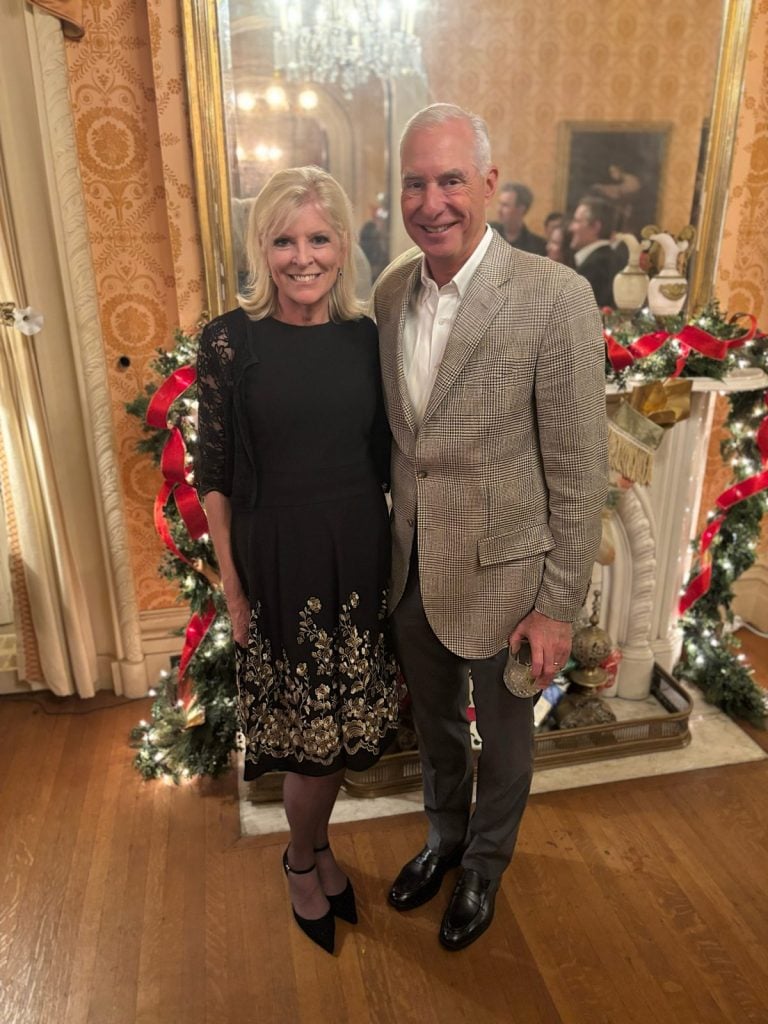 A woman in a black dress and a man in a light-colored blazer stand smiling together in front of a decorated fireplace with garlands and red bows in an elegant room.