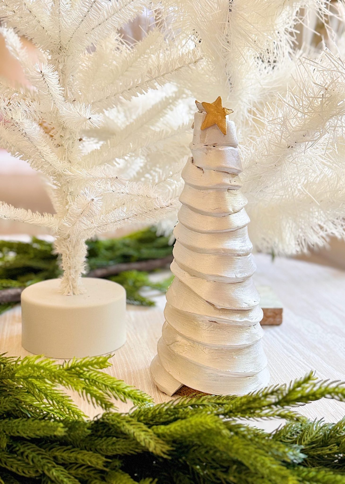 A small, white, spiral Christmas tree with a star on top sits on a light wooden surface beside artificial white trees and green fir branches.