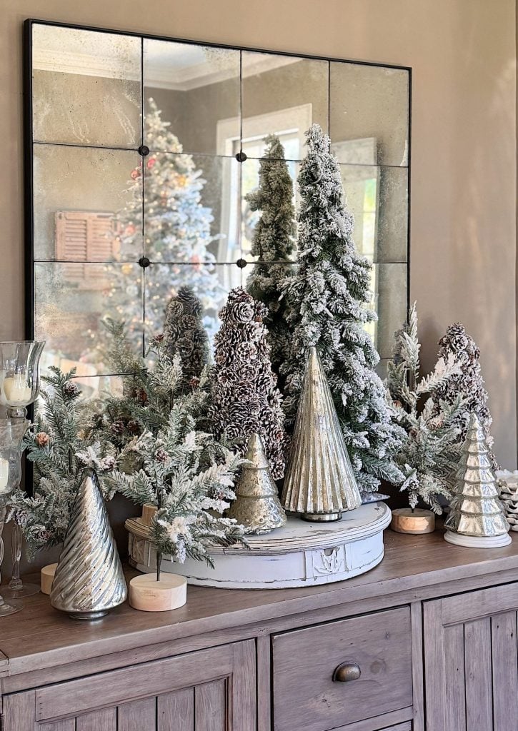 A wooden sideboard displays snowy artificial trees, metallic cone decorations, and candles, with a large mirror behind reflecting a Christmas tree.