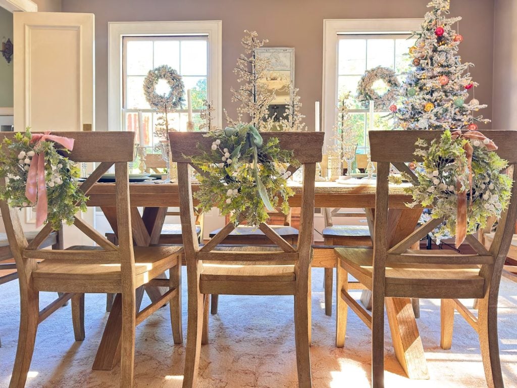 A dining room with a wooden table and chairs, decorated with wreaths and holiday ornaments, with sunlight streaming through two windows and a Christmas tree in the corner.