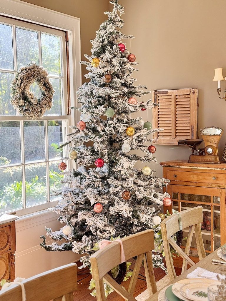 A flocked Christmas tree decorated with colored ornaments stands by a window with a wreath in a sunlit, rustic dining room.