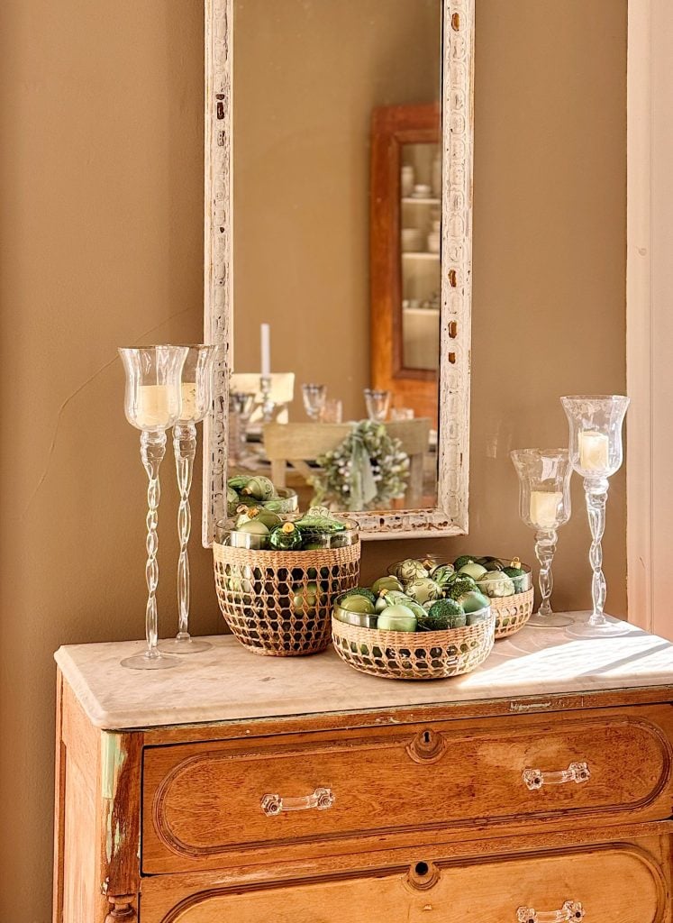 A wooden dresser with a marble top holds three baskets of green ornaments and four glass candle holders, with a distressed mirror hanging above.