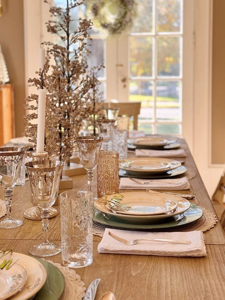 A wooden dining table is set for a meal with layered plates, silver cutlery, glassware, white napkins, and decorative centerpieces featuring branches and candles.