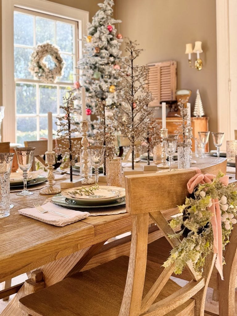 A wooden dining table set with plates, glasses, candles, and centerpiece trees; a decorated Christmas tree and wreath are visible by the window in a sunlit room.