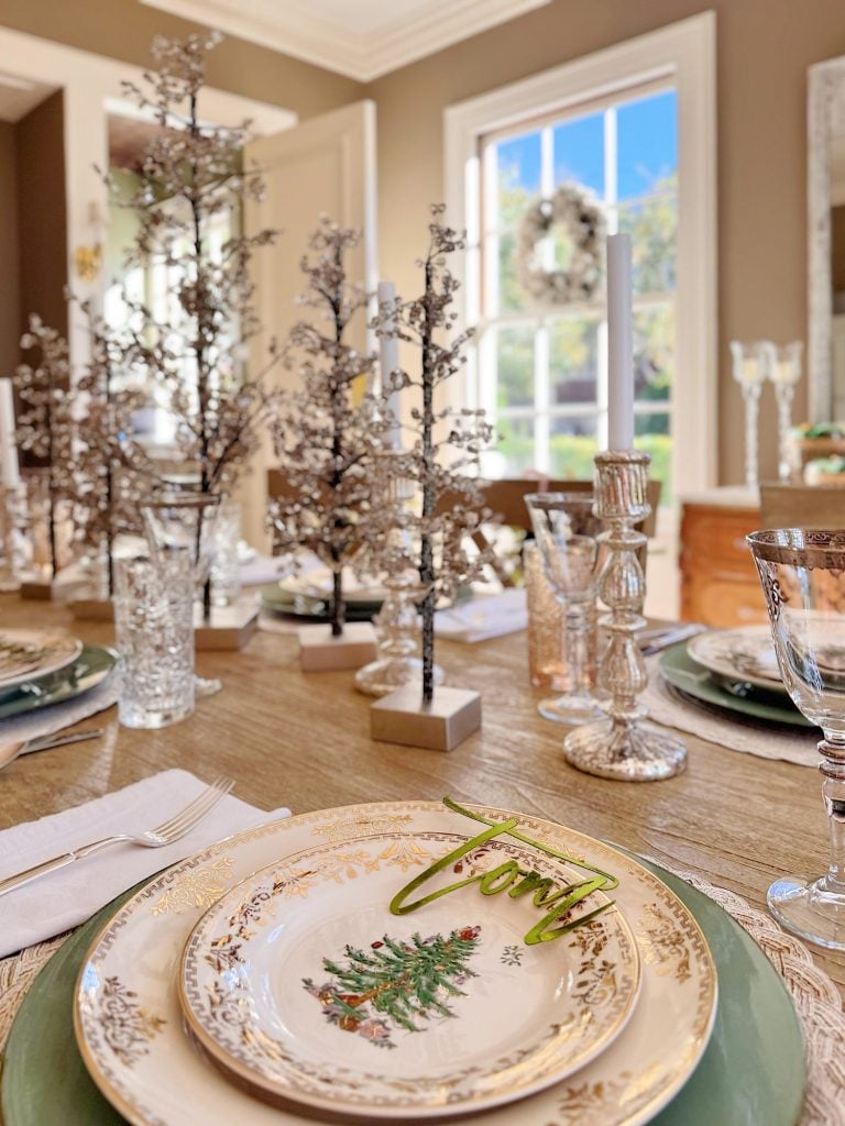 A festive dining table set with Christmas-themed plates, glassware, silverware, crystal candle holders, and decorative mini trees, with natural light coming through a window.