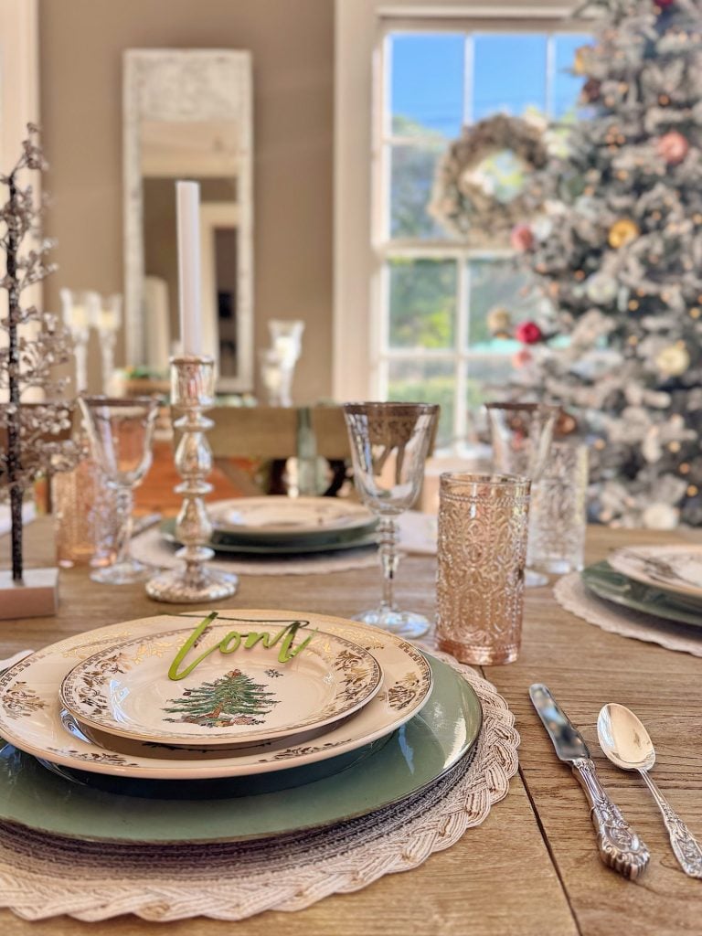 A dining table is set with holiday-themed plates, glassware, and silverware. A decorated Christmas tree and wreath are visible in the background by a large window.