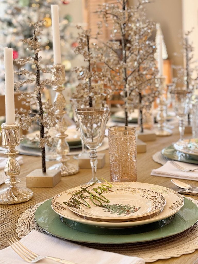 A festive holiday table is set with vintage plates, green chargers, crystal glasses, silver candlesticks, and decorative trees, with a Christmas tree visible in the background.