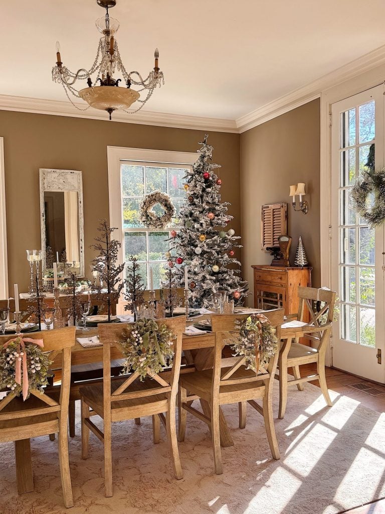 A dining room decorated for Christmas with a tree, wreaths, and festive centerpieces on a wooden table, sunlit through large windows.