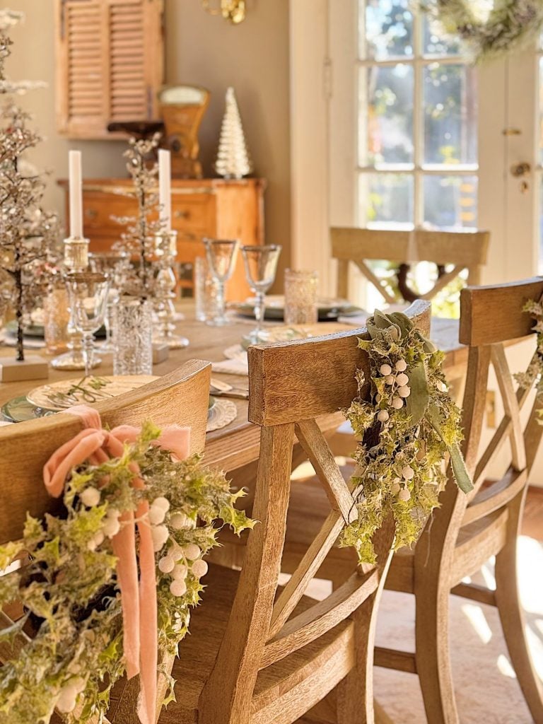 A sunlit dining room with a wooden table set for a meal, decorated with glassware, candles, and wreaths on the chairs.