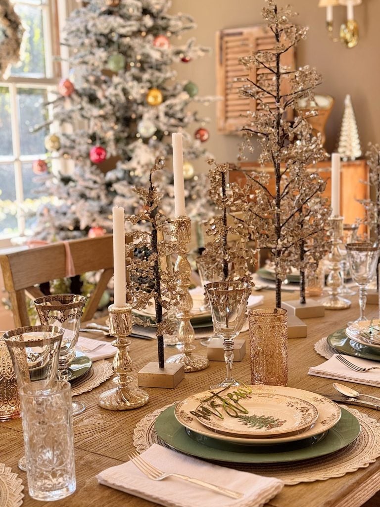 A decorated dining table set with plates, glasses, candlesticks, and small ornamental trees; a Christmas tree stands in the background.