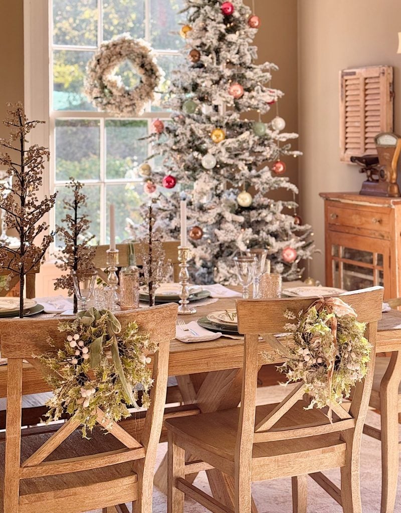 A dining room with a wooden table set for a meal, decorated chairs, a frosted Christmas tree with ornaments, and a wreath on the window in the background.