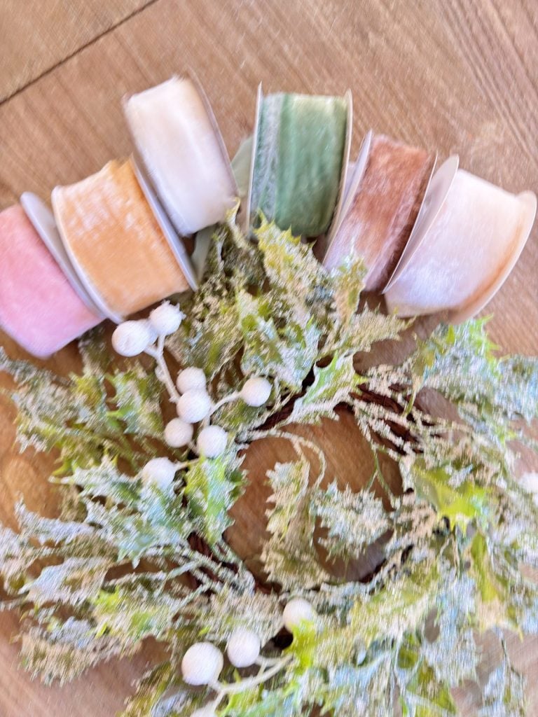 A frosted artificial wreath with white berries is displayed on a wooden surface next to five rolls of pastel-colored ribbon.