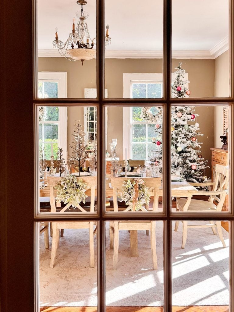 A dining room decorated for Christmas with a set table, wooden chairs, a chandelier, and a Christmas tree near two sunlit windows, viewed through glass-paneled doors.