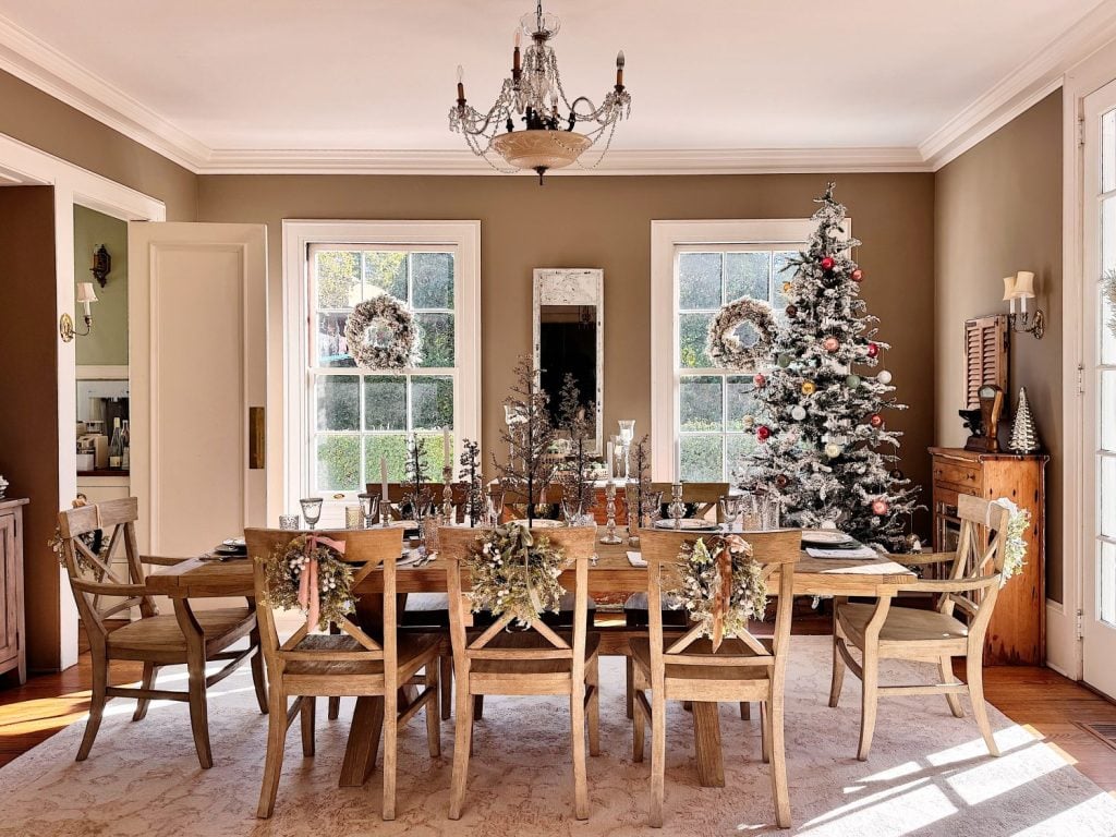 A dining room with a long wooden table set for a meal, decorated with festive wreaths and a Christmas tree near large windows.