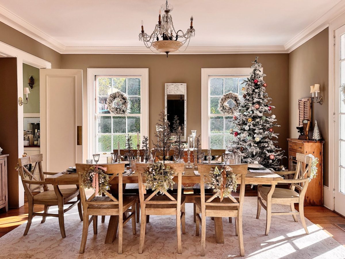 A dining room with a long wooden table set for a meal, decorated with festive wreaths and a Christmas tree near large windows.