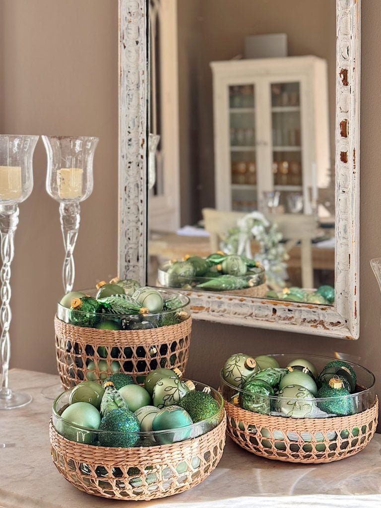 Three wicker baskets filled with green and gold Christmas ornaments sit on a table beneath a distressed white mirror. Two glass candle holders are placed nearby. A cabinet is reflected in the mirror.
