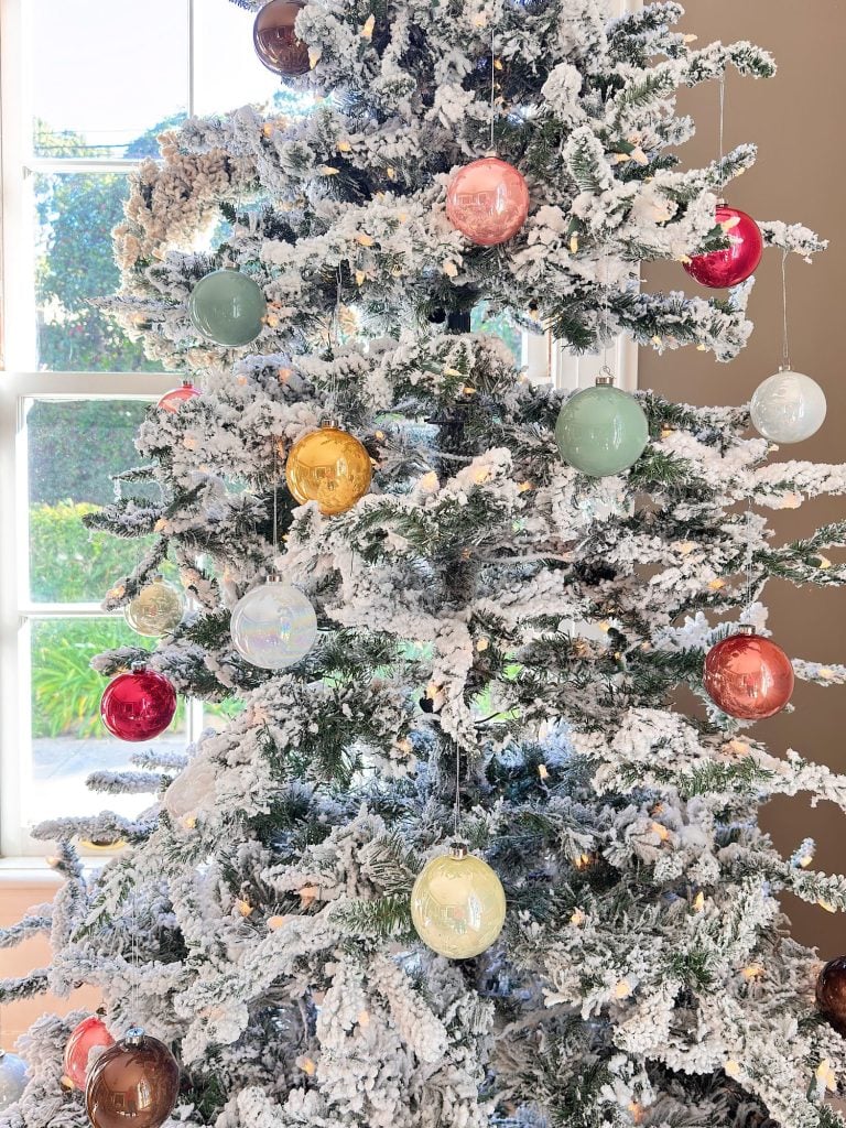 A flocked Christmas tree decorated with assorted colorful ornaments stands near a bright window in a sunlit room.