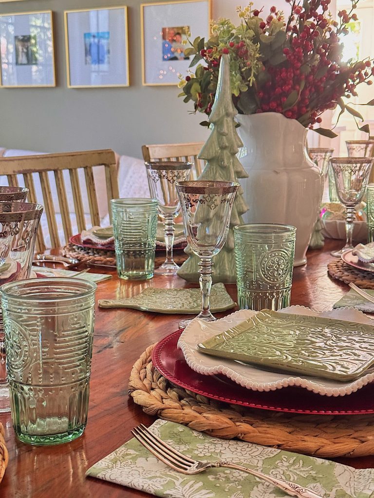 A dining table set with green and clear glassware, patterned plates, napkins, and a white vase with red berries and greenery as a centerpiece. Framed art hangs on the wall in the background.