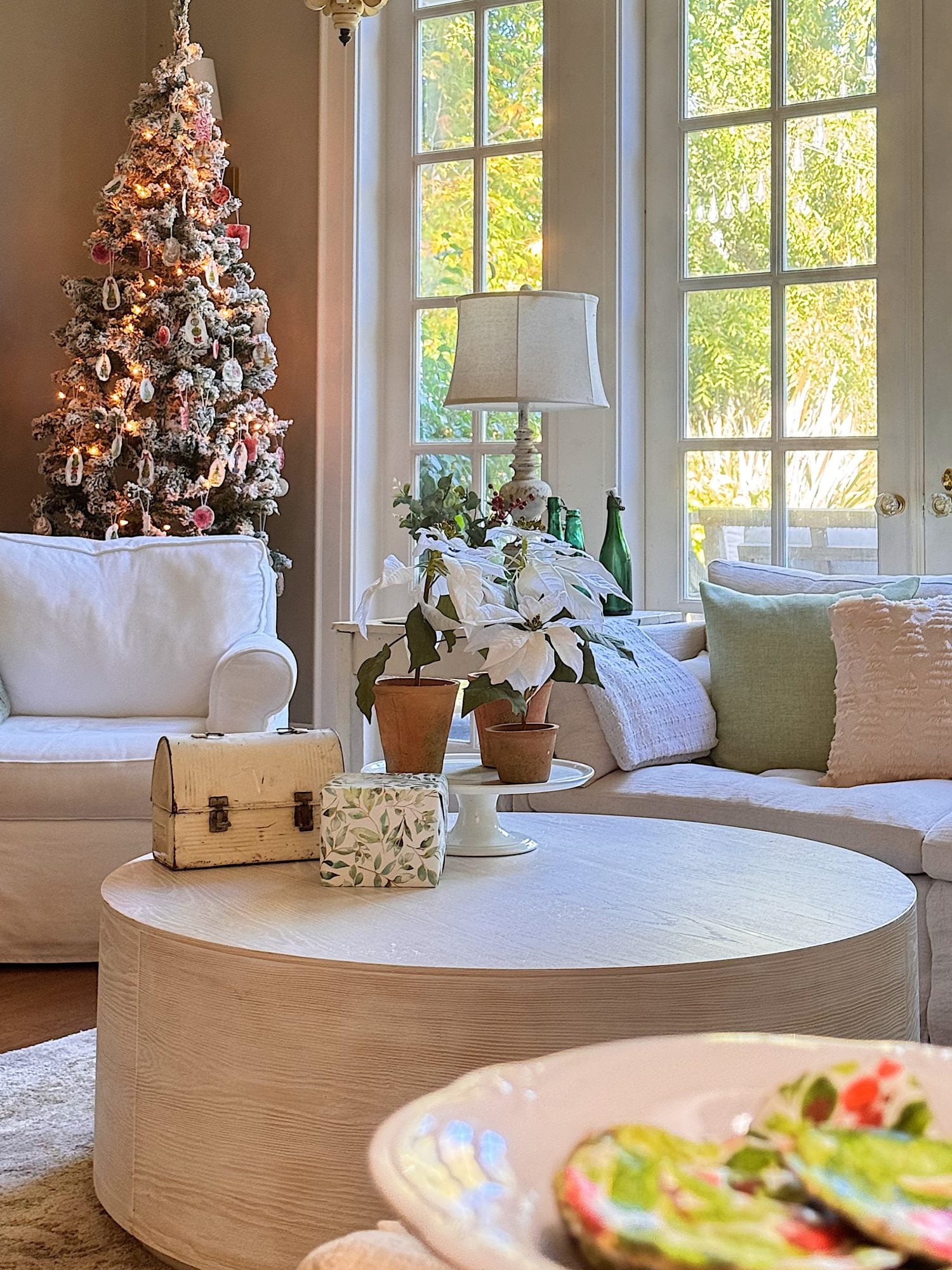 A cozy living room with a decorated Christmas tree, white sofas, a round coffee table with wrapped gifts and potted plants, and sunlight streaming through large windows.