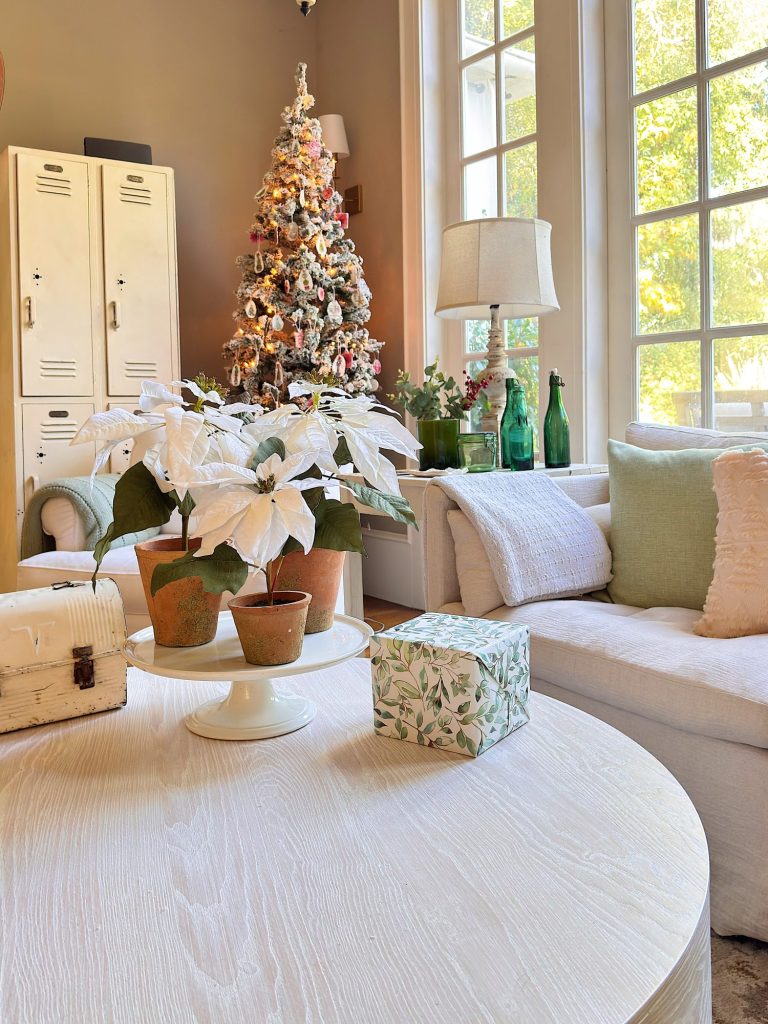 A cozy living room with a decorated Christmas tree, white poinsettias on a round coffee table, a sofa with light pillows, and several green bottles near large windows.