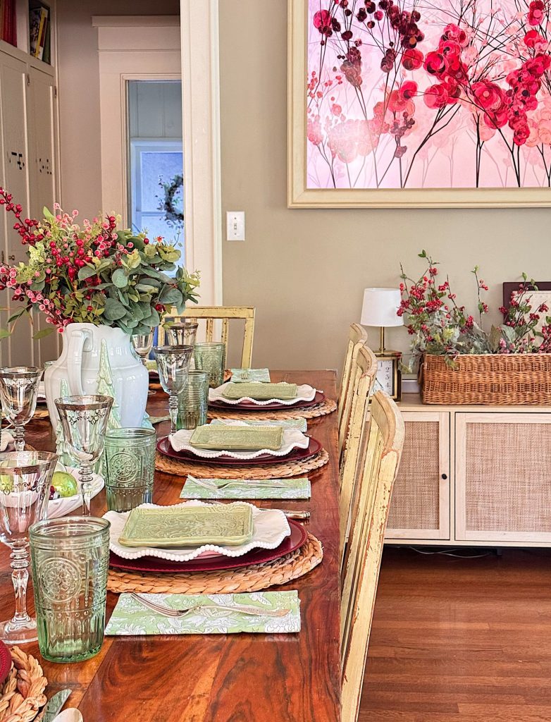 A wooden dining table set with green glassware, layered plates, and floral decor; a large pink floral painting and more flowers are visible in the background.