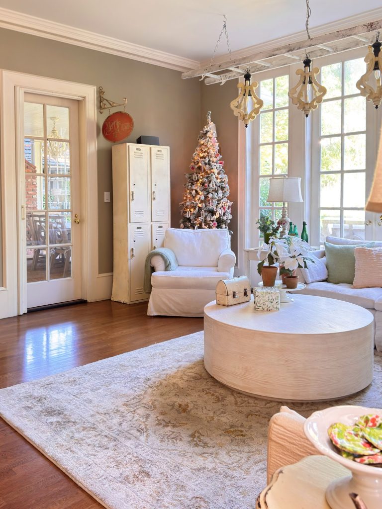 Bright living room with a decorated Christmas tree, white armchair, round coffee table, lockers, large windows, and neutral-toned furnishings.