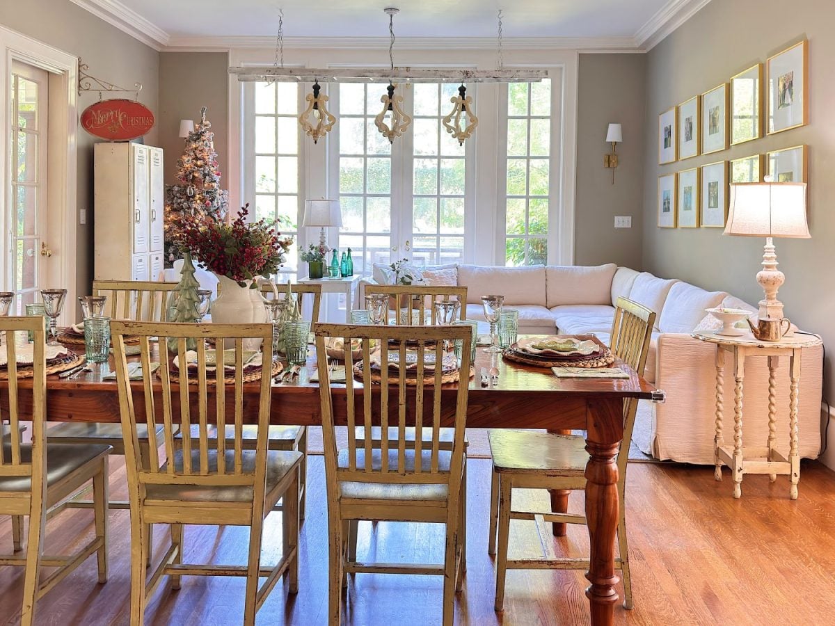 A dining and living area with a wooden table set for a meal, light wood chairs, a beige sectional sofa, framed wall art, and large windows letting in natural light.