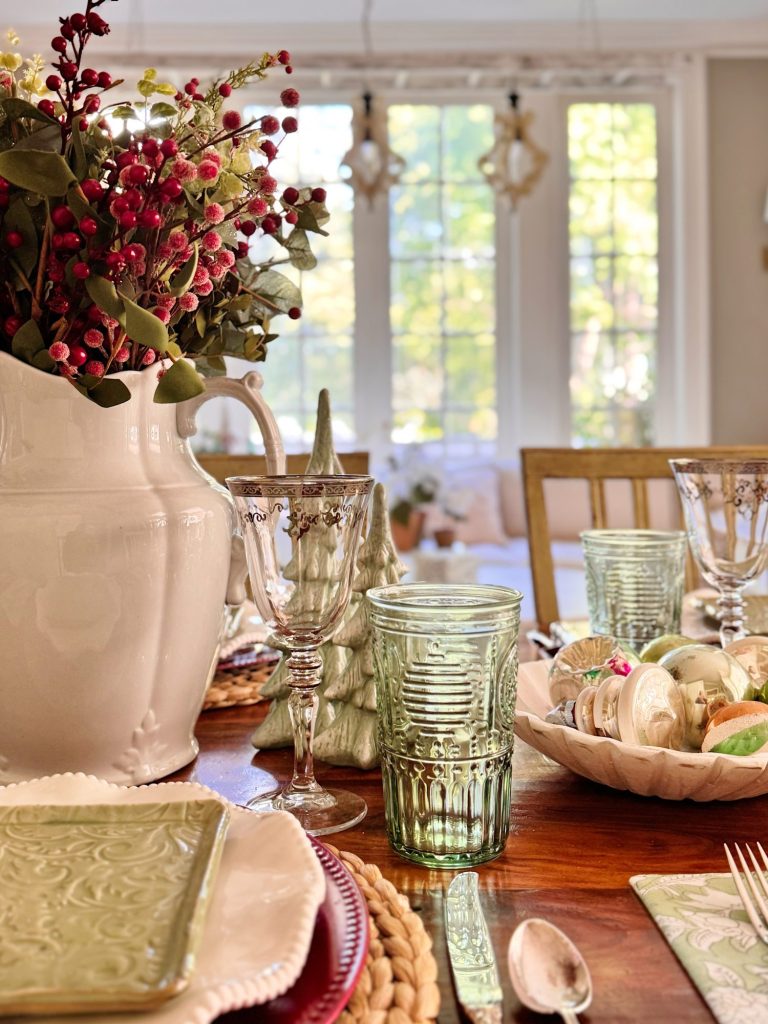 A dining table set with glassware, plates, and a large white vase with red berries and greenery; sunlight streams through large windows in the background.