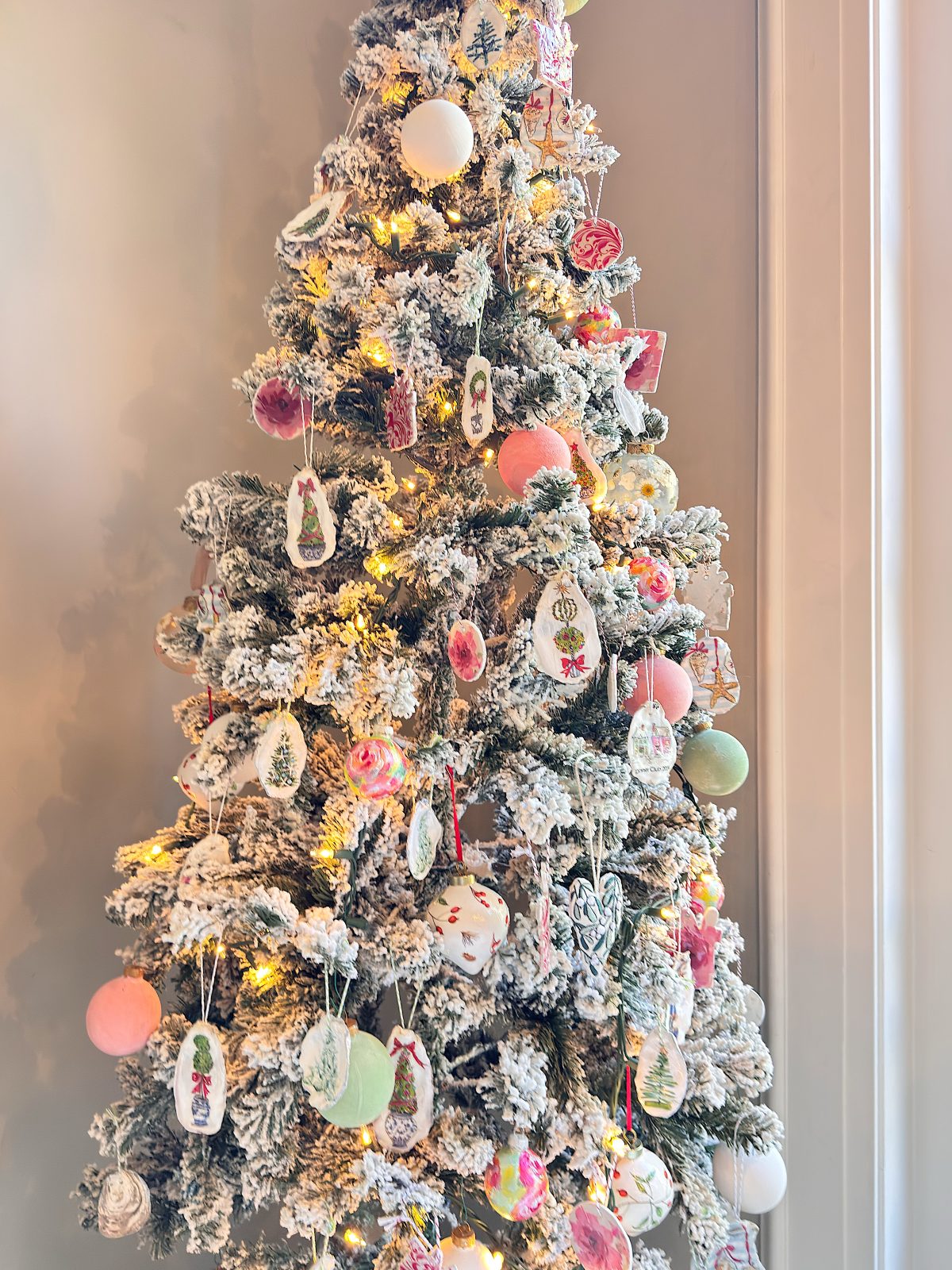 A flocked Christmas tree, adorned with assorted colorful ornaments and warm white string lights, brings festive Christmas decor to the space as it stands by a window.