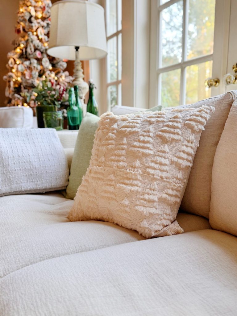 A close-up of a beige sofa with textured and smooth pillows, set in a living room with large windows and a decorated Christmas tree in the background.
