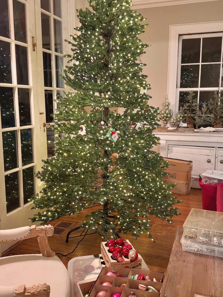 A partially decorated Christmas tree with lights stands in a living room, surrounded by boxes and containers of ornaments and decorations.
