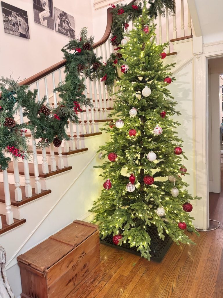 A decorated Christmas tree with red and white ornaments stands beside a staircase adorned with garland and pinecones in a home with wooden floors.