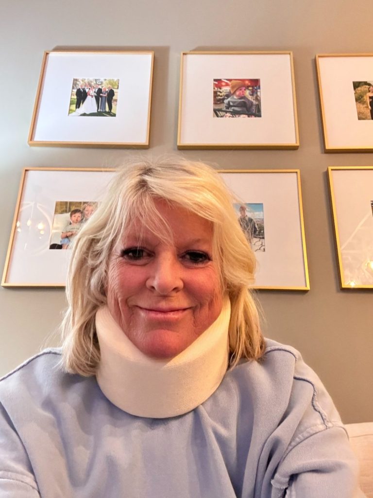 A woman with blonde hair wearing a neck brace and a light blue top sits indoors; framed photos are visible on the wall behind her.