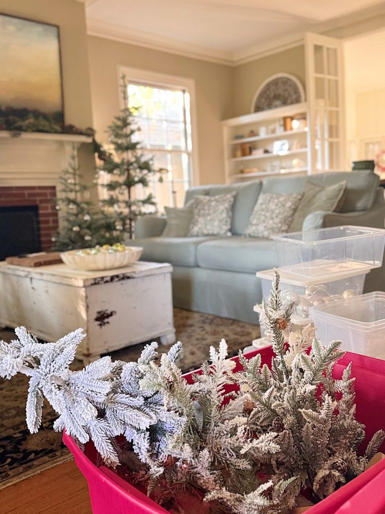 A living room with a sofa, coffee table, and small Christmas trees; a red bin in the foreground holds artificial snowy branches.