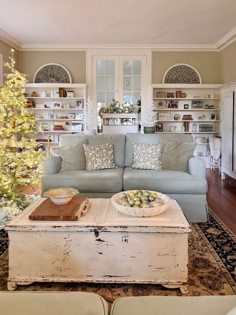 A cozy living room with a light blue sofa, a distressed white coffee table holding ornaments, a decorated Christmas tree, and built-in shelves with books and decor.